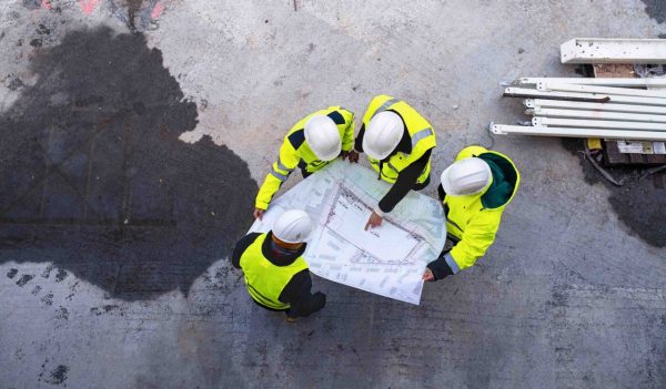 A top view of group of engineers with blueprints standing on construction site.