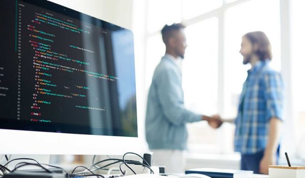 Two males shaking hands in background, focus on computer monitor with programming code and messy cables on table