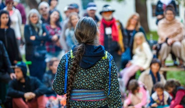 Woman with platted hair and glasses is giving a public speech or conference at the park in front of a blurry crowd of 50 people.