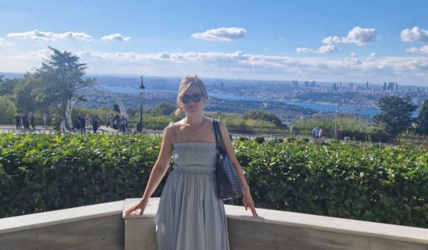 Lucy standing on a balcony in a part area with river and city skyline in the distance on a sunny day.