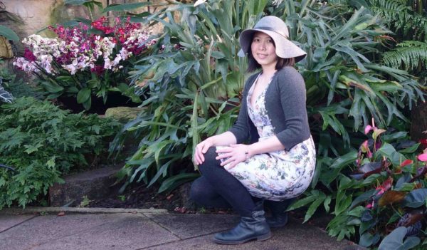 Sabrina Tang crouched in front of a flower garden, wearing a floppy sun hat.