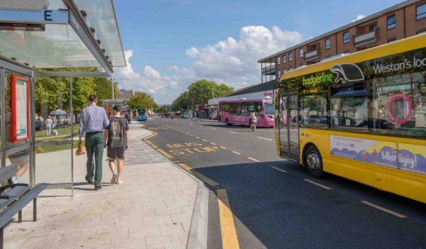 Bus stop with pedestrians on the pavement and a bus driving past on a sunny day