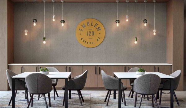 Interior resident common area showing two tables with seating for 4 with a feature wall in the background with light pendantds and a wall feature with the  building name - Emblem 120.