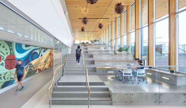 Interior view of a hallway with a long mural, with a staircase in the middle and benches / sitting areas alongside the stairs with large windows.