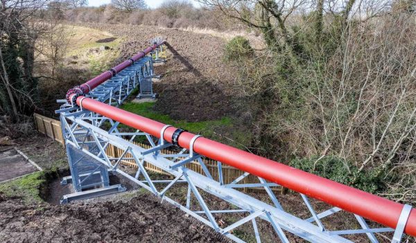 Long red pipeline elevated on metal supports running through a rural landscape with trees and a wooden fence nearby